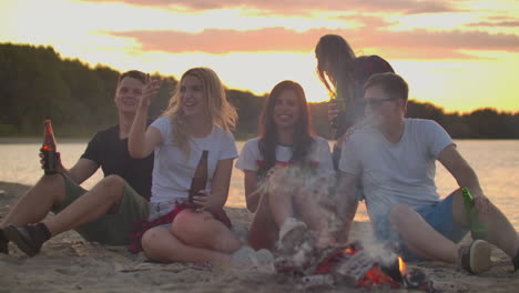 The-young-people-are-sitting-in-around-bonfire-on-the-river-coast.-They-are-laughing-and-enjoying-beer-at-sunset-in-summer-evening.